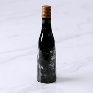 May include: A tall, slender bottle with a glossy black body featuring subtle, swirling white marble-like patterns. The bottle is topped with a cylindrical, ribbed wooden stopper that has a warm, light brown tone and a smooth, polished finish.