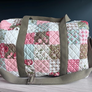 May include: A quilted duffel bag featuring a patchwork design of various floral, plaid, and polka dot patterns in shades of pink, white, brown, and light blue. The bag includes olive green fabric handles and an adjustable olive green shoulder strap.