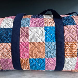 May include: A quilted duffel bag featuring a patchwork design of various squares in shades of pink, blue, orange, and white. The fabric squares display patterns including rainbows, stars, and geometric shapes. Two dark navy blue fabric straps are attached.