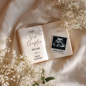 Pode incluir: Um livro aberto com o texto "OUR New Chapter BEGINS" e "BABY HARPER" em escrita elegante. Uma foto de ultrassom com o texto "APRIL 2026" está ao lado do livro. A cena é decorada com flores brancas em um fundo neutro.