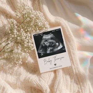 May include: A rectangular card featuring a black and white ultrasound image of a fetus, with the printed text Baby Thomson, a small heart icon, and the date DECEMBER 2026. The card is displayed alongside a sprig of white baby's breath flowers.