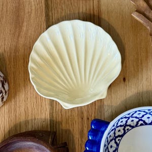 May include: A cream-colored ceramic dish shaped like a scallop shell. The dish features a glossy finish with distinct, radiating ridges that extend from the base to the scalloped edge, creating a textured, fan-like appearance for this small bowl.