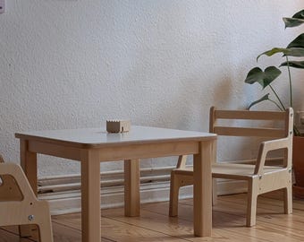 May include: A light wood children's table and two matching chairs. The square table has a light-colored top. Two framed art pieces hang on the wall. A colorful stacking toy and a small rug are on the wooden floor.