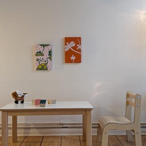 May include: A child's room scene featuring a small wooden table and chair set. The table has a white top and light wood legs. Two small framed art pieces hang on the wall. A toy animal and wooden blocks sit on the table.