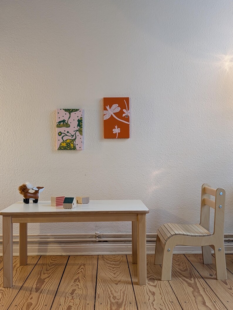 May include: A child's room scene featuring a small wooden table and chair set. The table has a white top and light wood legs. Two small framed art pieces hang on the wall. A toy animal and wooden blocks sit on the table.
