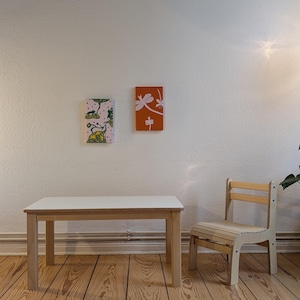 May include: A child's wooden table and chair set with a white tabletop and light wood legs. Two small framed artworks hang on the wall, one with a dragonfly design on an orange background and the other with a floral pattern.