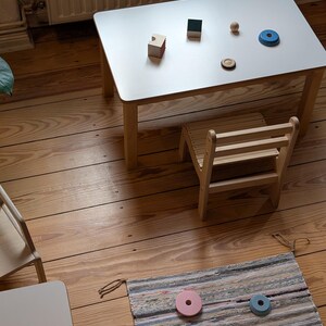 May include: A small, light-colored wooden table with a white top, accompanied by a matching chair. Various wooden toys, including blocks and rings, are arranged on the table. A colorful rug with wooden toys is on the floor. The table is approximately 24 inches long.