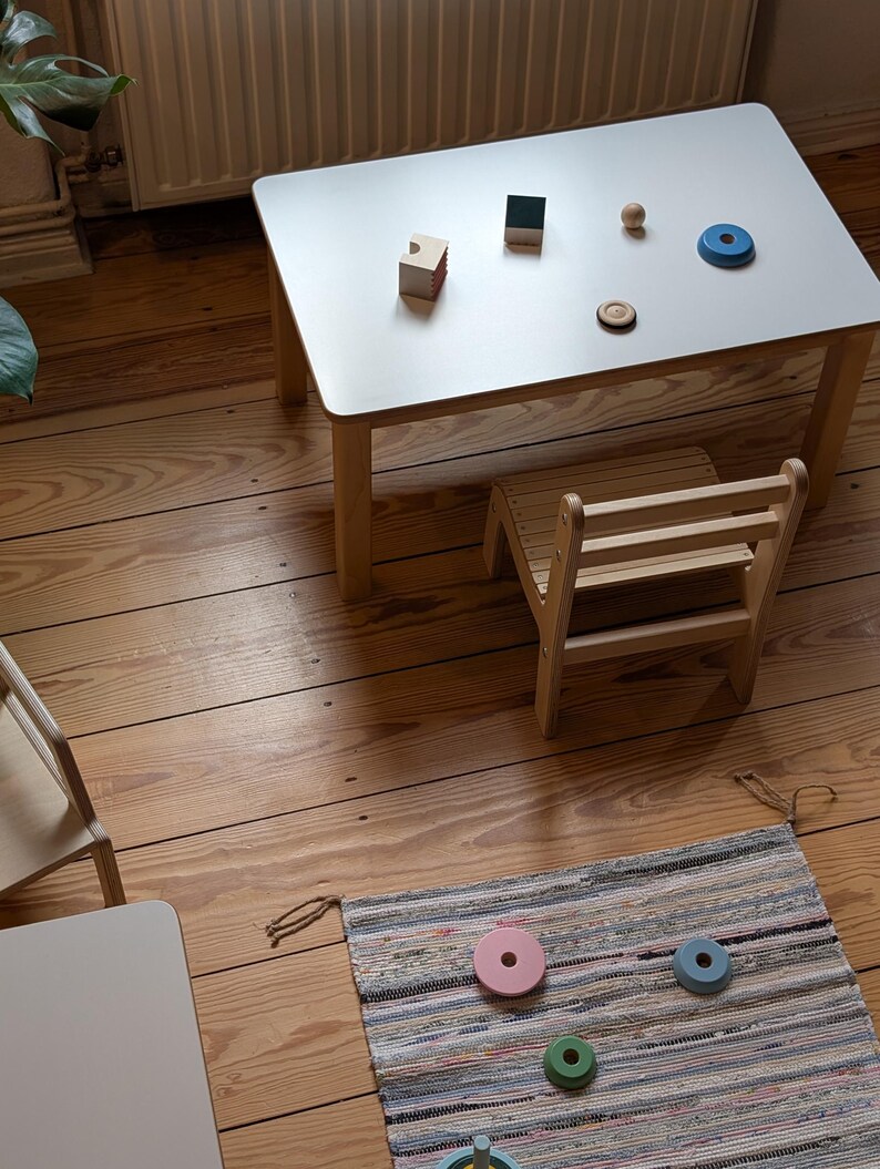 May include: A small, light-colored wooden table with a white top, accompanied by a matching chair. Various wooden toys, including blocks and rings, are arranged on the table. A colorful rug with wooden toys is on the floor. The table is approximately 24 inches long.