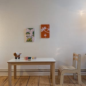 May include: A child's room scene featuring a small white table with wooden legs, a matching chair, and two framed art pieces on the wall. The artwork includes a dragonfly and a whimsical animal scene. A toy animal and wooden blocks are on the table.