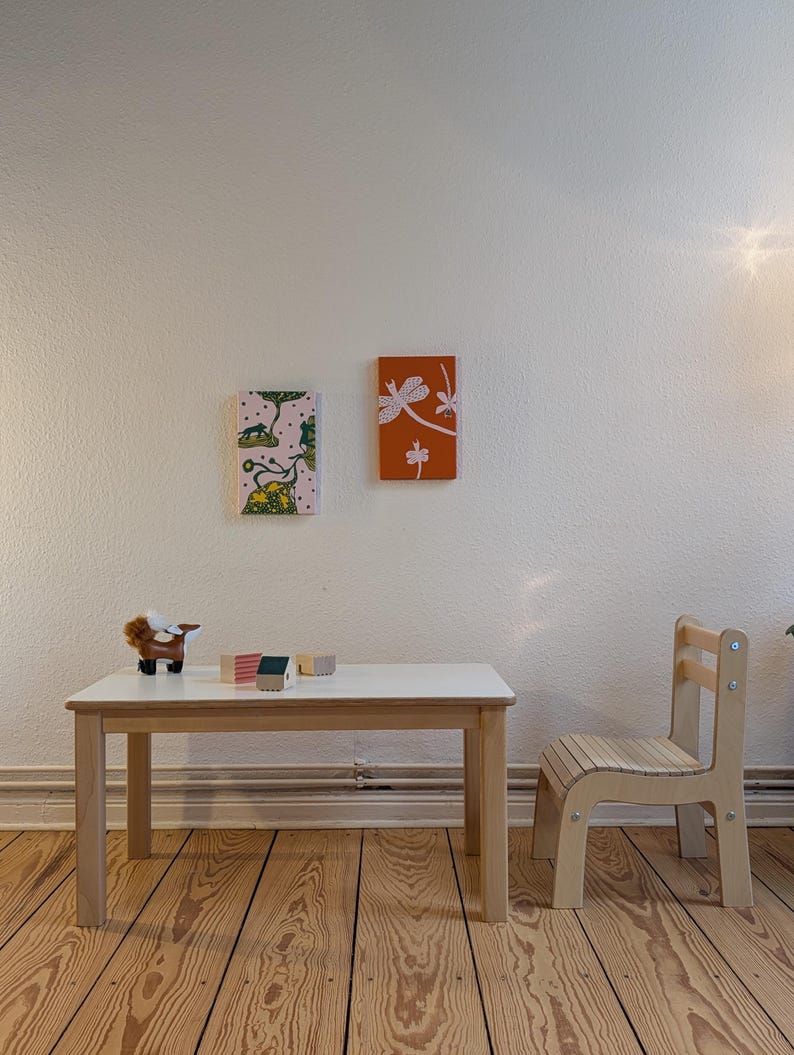 May include: A child's room scene featuring a small white table with wooden legs, a matching chair, and two framed art pieces on the wall. The artwork includes a dragonfly and a whimsical animal scene. A toy animal and wooden blocks are on the table.