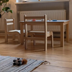 May include: A light-colored wooden children's table and two chairs set. The table has a rectangular top, and the chairs have a ladder back design. A small rug with a toy car is in the foreground.