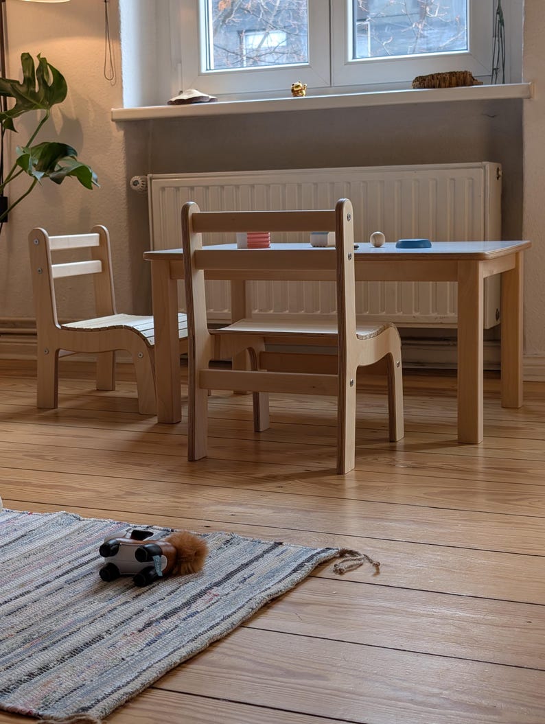 May include: A light-colored wooden children's table and two chairs set. The table has a rectangular top, and the chairs have a ladder back design. A small rug with a toy car is in the foreground.