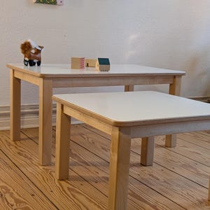 May include: Two light wood square tables with white tops, one larger than the other, are shown. A small wooden horse figurine and miniature wooden houses sit on the larger table. The tables are on a wood floor.