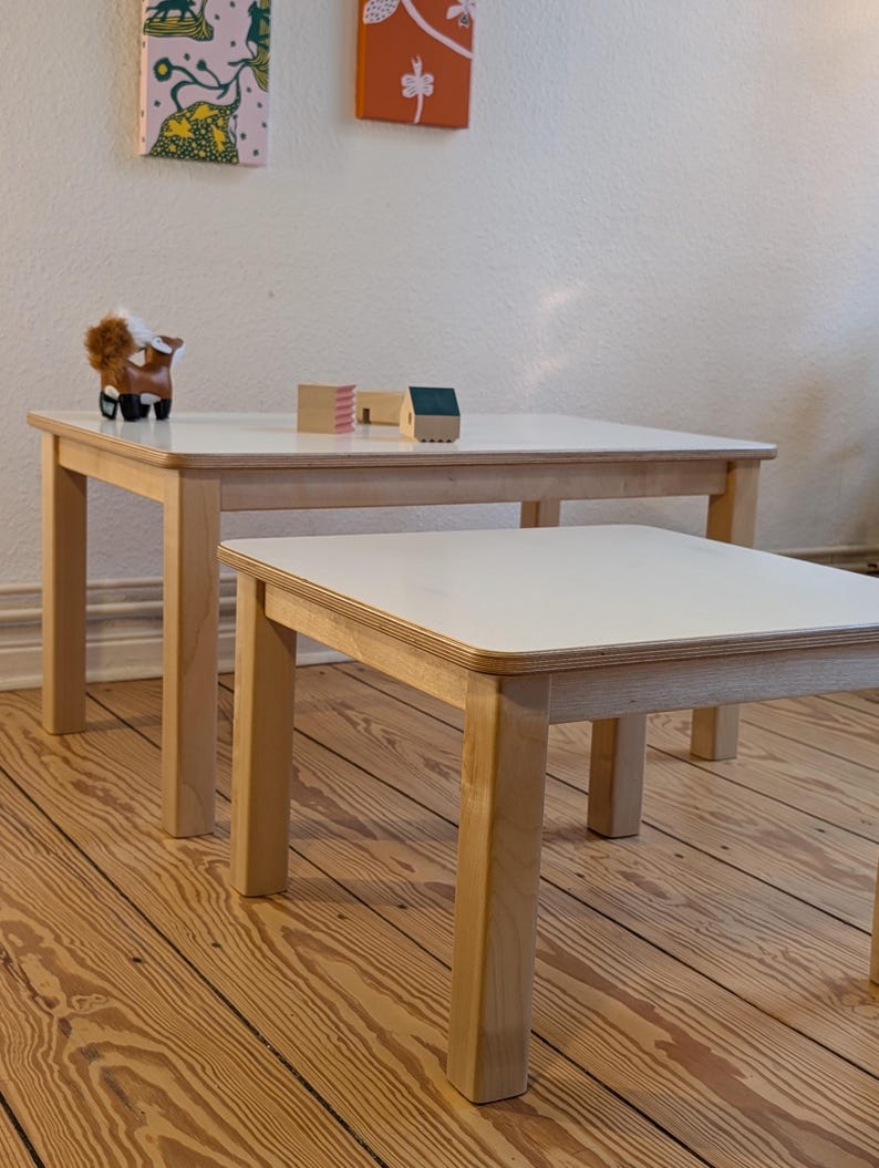 May include: Two light wood square tables with white tops, one larger than the other, are shown. A small wooden horse figurine and miniature wooden houses sit on the larger table. The tables are on a wood floor.
