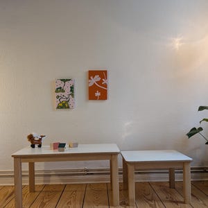 May include: Two wooden tables with white tops and light brown legs. A larger rectangular table is next to a smaller square table. Two small framed artworks hang on the wall above the tables. A toy animal and small wooden blocks are on the larger table.
