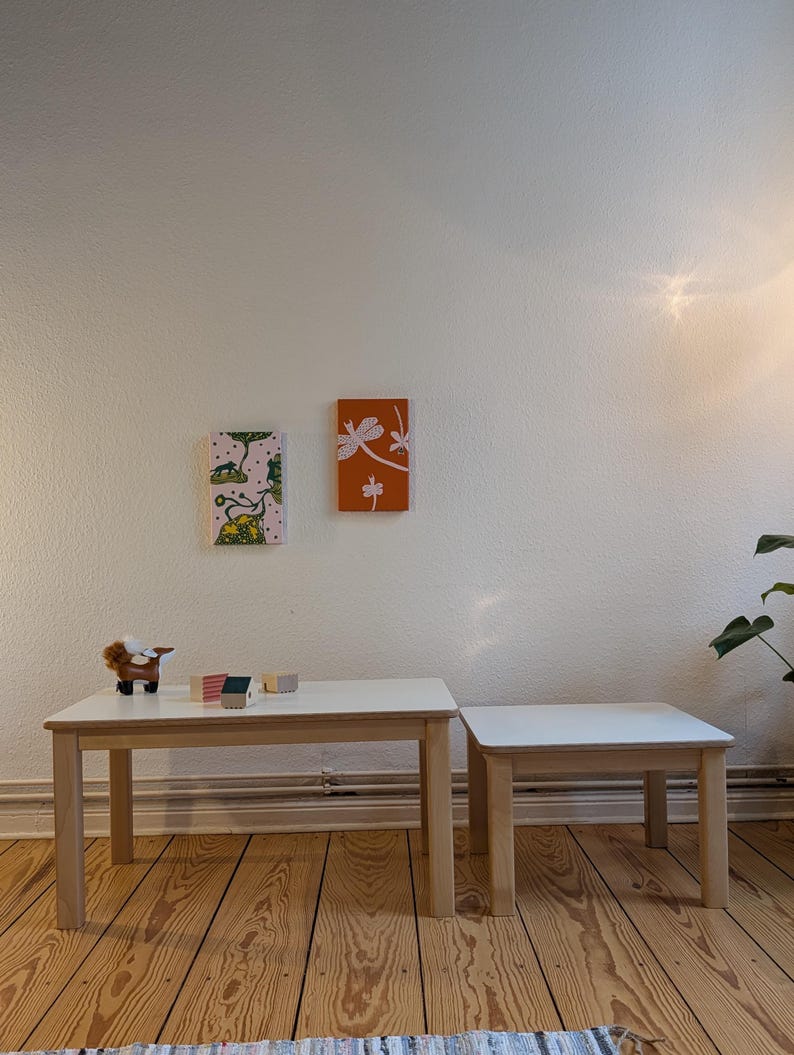 May include: Two wooden tables with white tops and light brown legs. A larger rectangular table is next to a smaller square table. Two small framed artworks hang on the wall above the tables. A toy animal and small wooden blocks are on the larger table.
