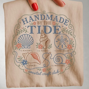 May include: A beige t-shirt featuring a blue and peach embroidered-style graphic that reads "HANDMADE BY THE TIDE" and "coastal craft club". The design includes illustrations of a scallop, turret, sand dollar, conch, and moon shell with floral accents.