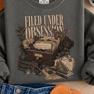 Op de afbeelding: Een antracietgrijze sweater met ronde hals en een grafische print met de tekst FILED UNDER OBSESSION. Het ontwerp toont een houten archiefkast, een nachtvlinder, een roos, vintage brieven, boeken, een vulpen en een bril met ronde glazen.