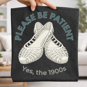May include: A black tote bag featuring a graphic of white, vintage-style jelly shoes. The text "PLEASE BE PATIENT" arches above the shoes, with "Yes, the 1900s" below. The bag is held by a hand.
