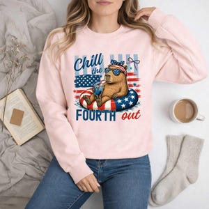 May include: A light pink sweatshirt featuring a capybara wearing sunglasses and a headband, floating in a patriotic inner tube. The text "Chill the Fourth out" is above and below the capybara, with an American flag design.