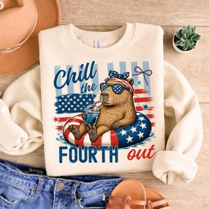 May include: Cream-colored sweatshirt with a graphic of a capybara wearing sunglasses and a patriotic headband, relaxing in a pool float. The text "Chill the Fourth Out" is displayed in blue. The design incorporates American flag elements.