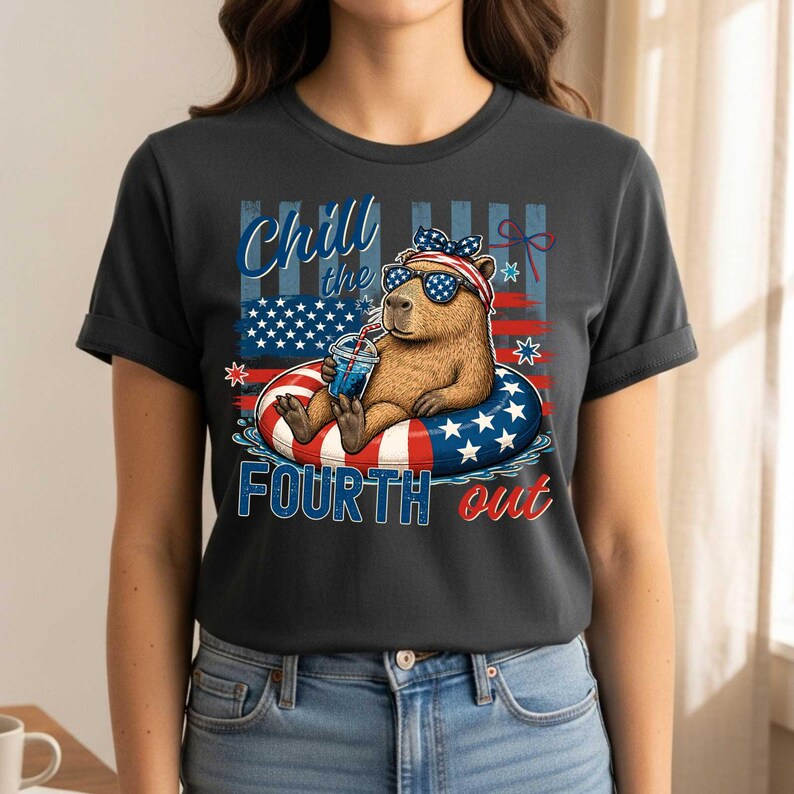 May include: Dark gray t-shirt featuring a capybara wearing sunglasses and a headband with an American flag design, floating on a red, white, and blue inner tube. The shirt has the text "Chill the Fourth out".