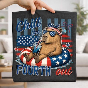May include: Dark gray tote bag featuring a capybara wearing sunglasses and a patriotic headband, floating in a red, white, and blue inner tube. The bag has the text "Chill the Fourth out" and an American flag design.