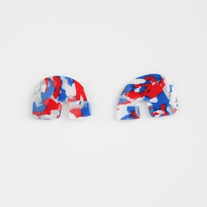 Op de afbeelding: Een paar boogvormige oorbellen met een gespikkeld patroon van rood, wit en blauw. De oorbellen zijn gemaakt van een glanzend materiaal en hebben een uitsparing in het midden, die op een regenboog lijkt.