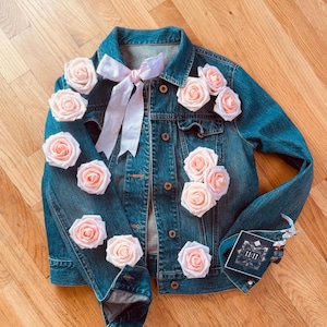 May include: A denim jacket adorned with light pink roses and a matching bow. The jacket features a classic button-up design and is decorated with multiple fabric roses. A small card with the text "11:11" is attached to the jacket.
