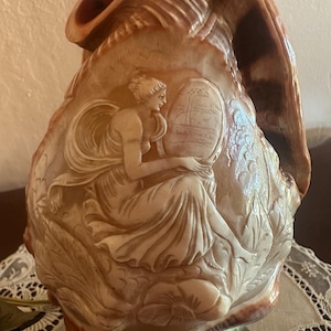 Puede incluir: Una gran concha marina pulida de color marrón y crema tallada con un relieve detallado de una figura sentada que sostiene un espejo ovalado. La concha presenta intrincados patrones florales y de follaje alrededor de la figura central, sobre una base metálica.