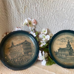 May include: Two round metal trays with dark blue scalloped edges and cream-colored centers. Each tray features a detailed sepia-toned illustration of a historic building with architectural columns and figures in the foreground, set against a white background.