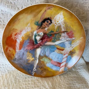 Puede incluir: Plato decorativo con una bailarina en movimiento. El plato tiene un borde dorado y una pintura vibrante con tonos rojos, azules y amarillos. La bailarina lleva una diadema floral roja y un tutú, y está de puntillas.