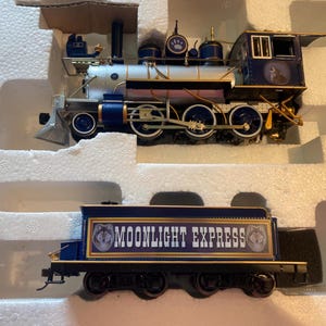 Puede incluir: Un modelo de locomotora de vapor y ténder con un esquema de color azul oscuro y plateado con detalles dorados. El ténder muestra el texto MOONLIGHT EXPRESS en letras blancas, flanqueado por gráficos de cabezas de lobo sobre fondo azul.