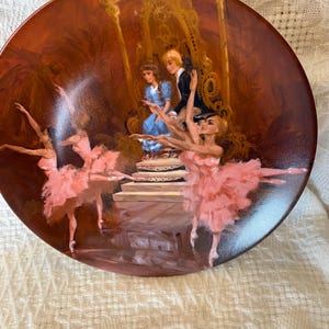 Puede incluir: Plato decorativo con una escena pintada que presenta bailarinas de ballet con tutús rosas y una pareja con ropa formal. El plato tiene un fondo marrón con detalles dorados y un acabado brillante.