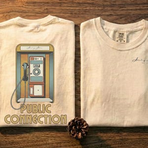 Könnte beinhalten: Zwei cremefarbene T-Shirts werden auf einer Holzoberfläche präsentiert, eines mit einer Retro-Telefonzellen-Grafik und den Worten "PUBLIC CONNECTION". Das andere Shirt hat ein kleines Logo. Vintage-Kassetten, Disketten und eine Kamera sind ebenfalls vorhanden.