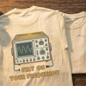 May include: Two cream-colored t-shirts with retro graphics and text. One shirt features an oscilloscope design with the words "STAY ON YOUR FREQUENCY." Other items include cameras, floppy disks, and cassette tapes, all on a wooden surface.