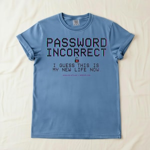 Op de afbeelding: Lichtblauw T-shirt met de tekst "PASSWORD INCORRECT" in gepixelde, 3D-letters. De tekst "I GUESS THIS IS MY NEW LIFE NOW" en "ANALOG ATLAS // ERROR LOG" staan er ook op. Het ontwerp bevat een slotpictogram.