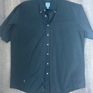 May include: A black short-sleeved button-down shirt with a button-down collar and a chest pocket. The shirt is hanging on a black hanger. The shirt has white buttons and a small blue tag on the collar.