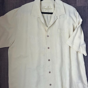 May include: A light yellow button-down shirt with short sleeves, a collar, and a chest pocket. The shirt has a subtle floral pattern and the brand name "Tommy Bahama" is embroidered on the collar.