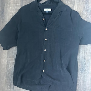 May include: A dark navy blue short-sleeve button-down shirt with a chest pocket. The shirt has a collared neckline and wooden buttons. The shirt is hanging on a black hanger.