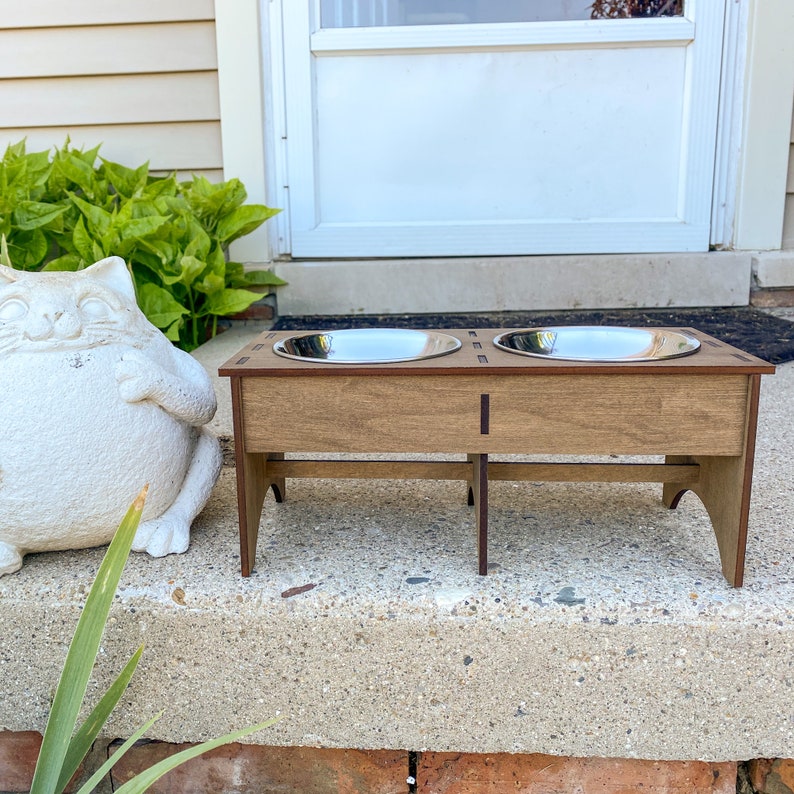 May include: A wooden pet food stand with two stainless steel bowls. The stand is brown and has a simple design. The bowls are elevated on the stand, making it easier for pets to eat.