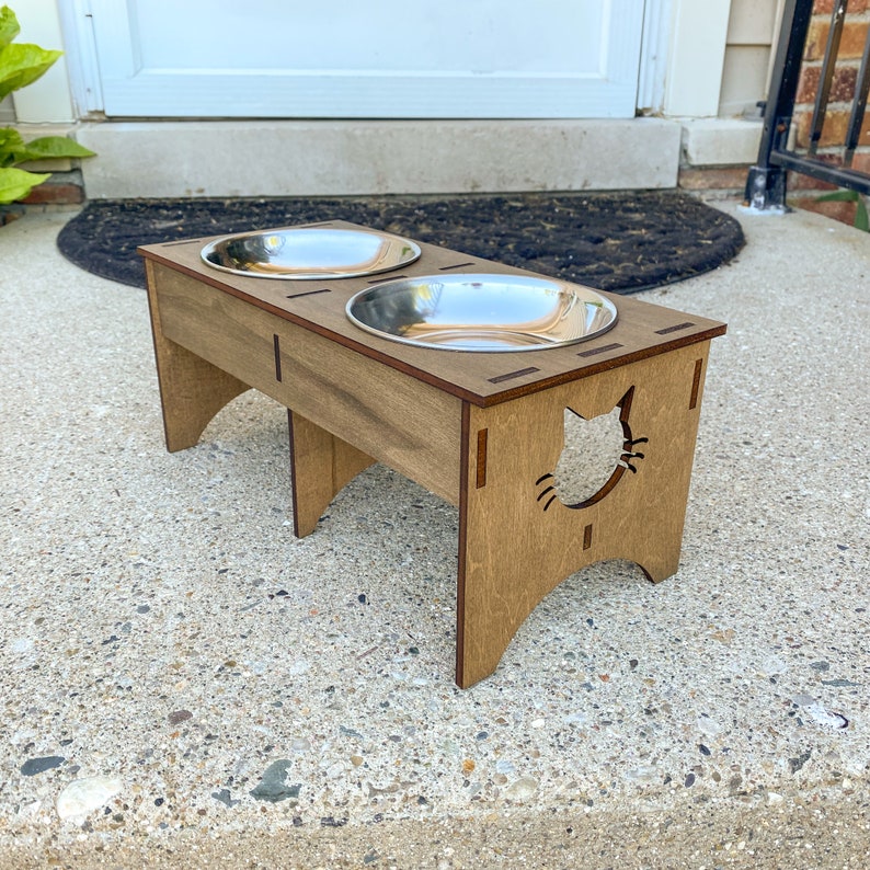 May include: A wooden elevated cat food and water bowl stand with two stainless steel bowls. The stand has a cat silhouette cutout.