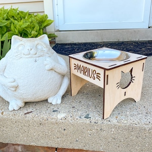 May include: A wooden cat food stand with a stainless steel bowl. The stand is engraved with the name "Marius" and a cat silhouette.