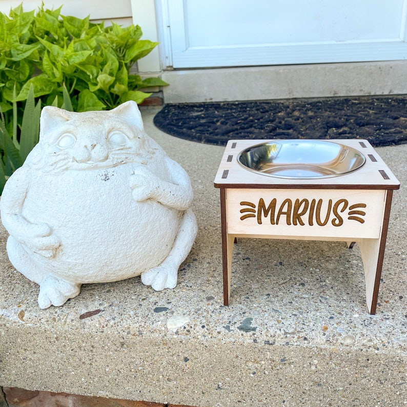 May include: A wooden pet food stand with a stainless steel bowl and the name "Marius" engraved on the front. The stand is elevated on four legs.