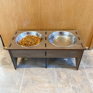 May include: A wooden pet food stand with two stainless steel bowls. One bowl is filled with dry pet food.