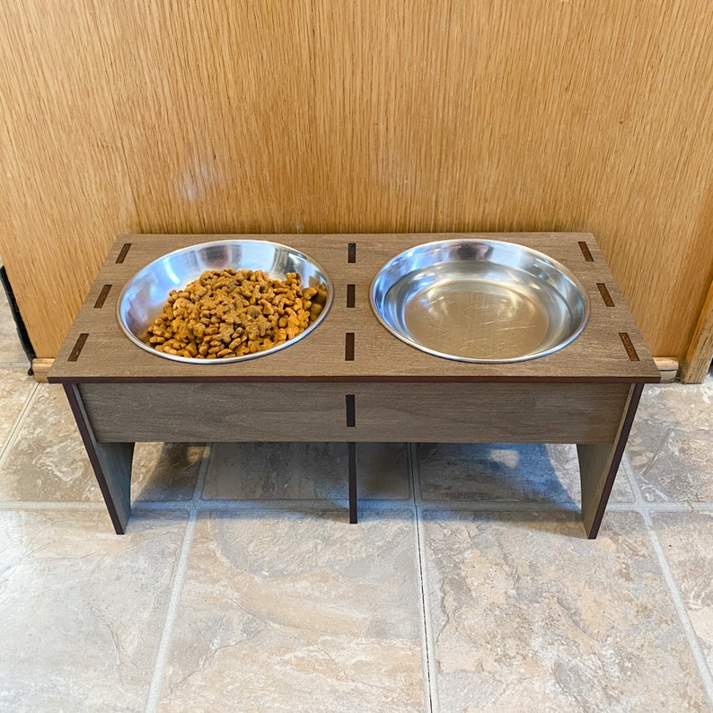 May include: A wooden pet food stand with two stainless steel bowls. One bowl is filled with dry pet food.