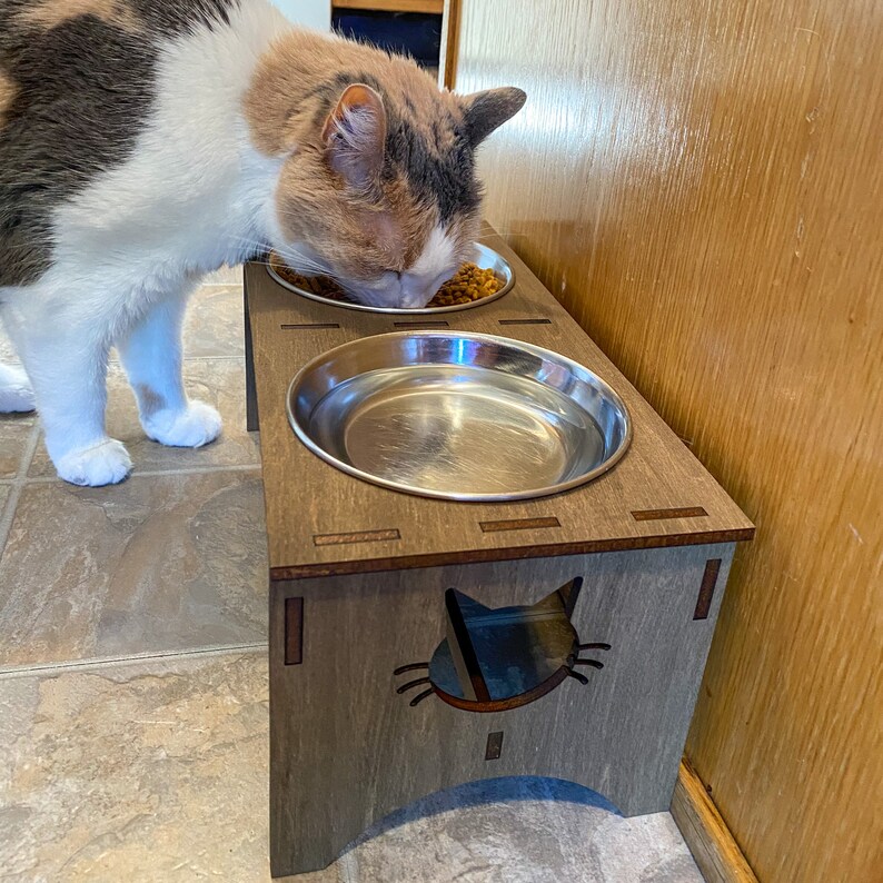 May include: A wooden cat food stand with two stainless steel bowls. The stand has a cat silhouette cutout and is stained a dark brown color. A cat is eating from one of the bowls.