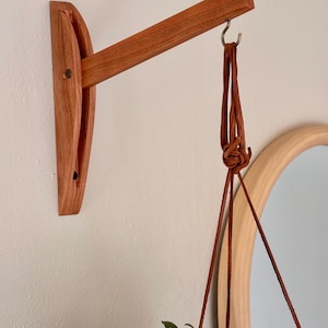 Puede incluir: Un colgador de plantas de madera con acabado natural está montado en una pared blanca. El colgador presenta un soporte curvo y un brazo horizontal con un gancho de metal. Una correa de cuero marrón sostiene una planta verde.