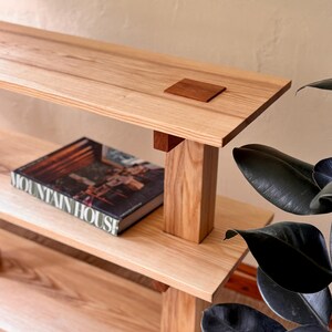 Op de afbeelding: Een lichtgekleurde houten plank met twee niveaus, ondersteund door verticale houten palen. Een kleine, vierkante, donkere houten onderzetter staat op de bovenste plank. Een boek met de titel "MOUNTAIN HOUSE" ligt op de onderste plank.