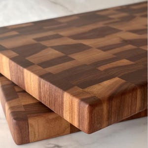Puede incluir: Dos tablas de cortar de testa. Las tablas son de madera marrón oscura y clara, con un patrón de tablero de ajedrez en la superficie superior. Los bordes son redondeados y lisos, y las tablas están apiladas una encima de la otra.